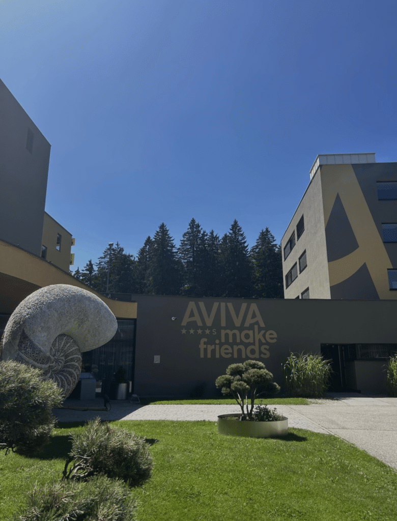 Haupteingang des Hotel AVIVA mit edlem goldenen Logo rechts neben der Tür und moderner anthrazitfarbener Fassade.